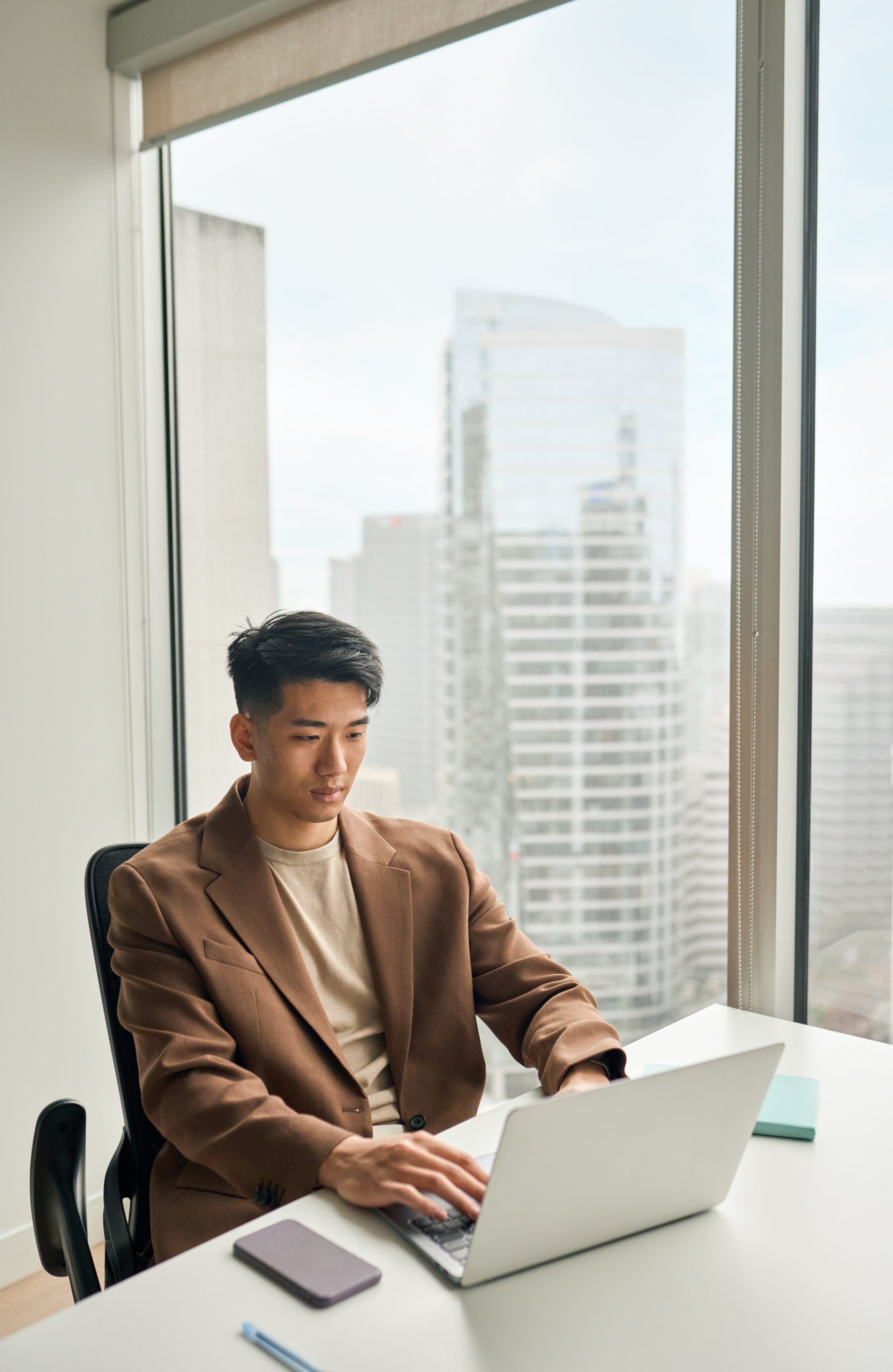 Man in blazer typing on laptop at office desk overlooking skyline.